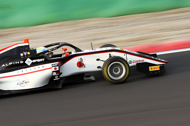 A Formula Regional race car with an Alpine Academy and Castrol livery speeds around a circuit. The driver, wearing a blue and yellow helmet, is visible in the cockpit, gripping the steering wheel. The car has a sleek aerodynamic design with sponsor logos, including Pirelli, and features black, white, and red accents. The blurred background and motion blur on the tires indicate high speed. The track surface is dry, with a red and white curb on the edge. Credit: Pastorelli Photography