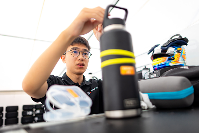 A young man wearing glasses and a black team polo shirt is working in a racing team environment. He is adjusting an item on a table, which has a black water bottle with yellow tape markings, a clear plastic container, and other racing gear. In the background, several racing helmets and equipment bags are visible. The setting appears to be inside a team tent or garage. Credit: FRECA / Dutch Photo Agency