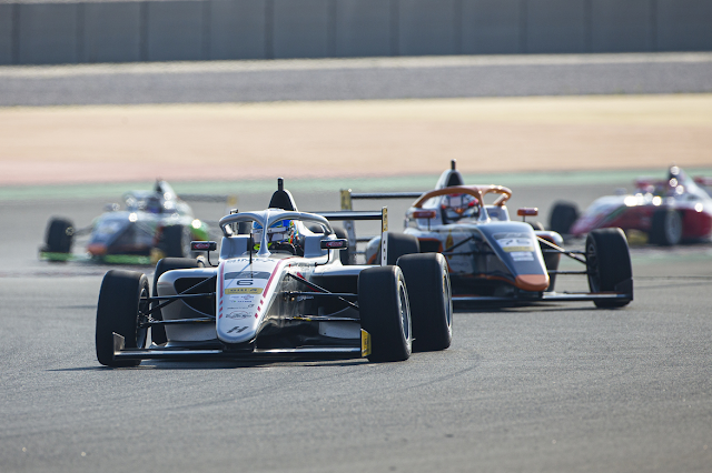A group of Formula 4race cars compete on a circuit, with the leading car number 6 being closely followed by others. The cars have aerodynamic front and rear wings, and the drivers are wearing helmets. The track surface is dry, and the background features barriers and a run-off area. The setting appears to be during a race or qualifying session in bright daylight. Credit: Formula 4 UAE