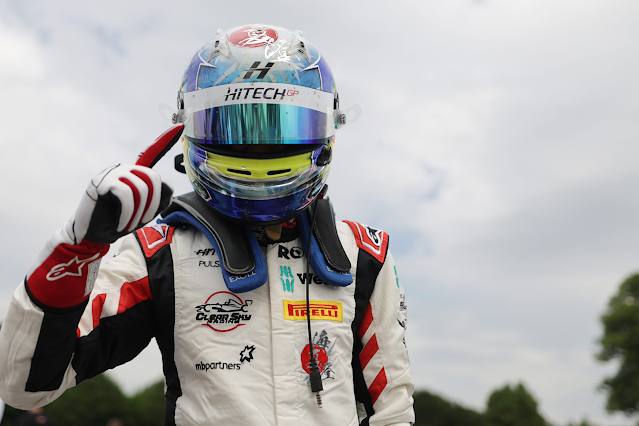 Alt text: A racing driver wearing a white and red racing suit and a reflective blue helmet with a "Hitech GP" visor strip celebrates by pointing their index finger upwards. The suit features multiple sponsor logos, including Pirelli and MB Partners. The driver is also wearing red and white racing gloves and a HANS device for safety. The background is slightly blurred with trees and a cloudy sky visible, indicating an outdoor racing circuit setting. Credit: F4 British Championship