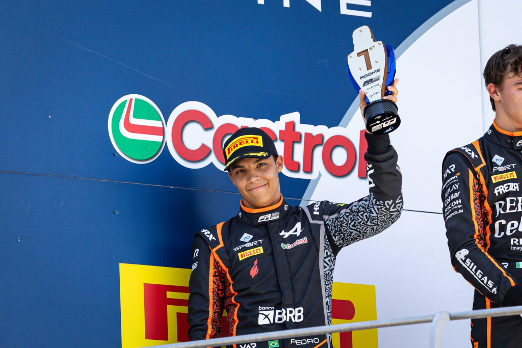 A young racing driver in a black and orange racing suit with Alpine, Castrol, and Pirelli branding smiles while holding up a "1st Rookie" trophy on the podium. He is wearing a Pirelli cap and has a confident expression. Another driver in a similar racing suit stands partially visible to the right. The backdrop features large Castrol and Pirelli logos, indicating an official motorsport event setting. Credit: FRECA / Dutch Photo Agency